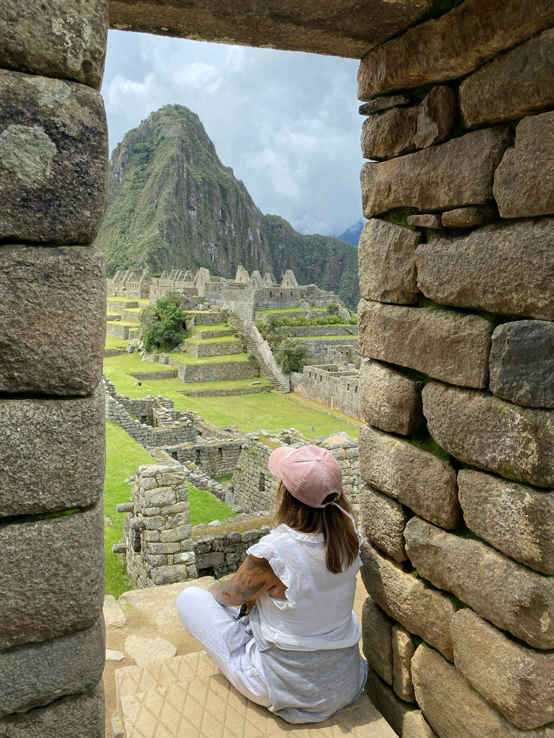 Woman in pink cap observes stunning Machu Picchu ruins through stone window.